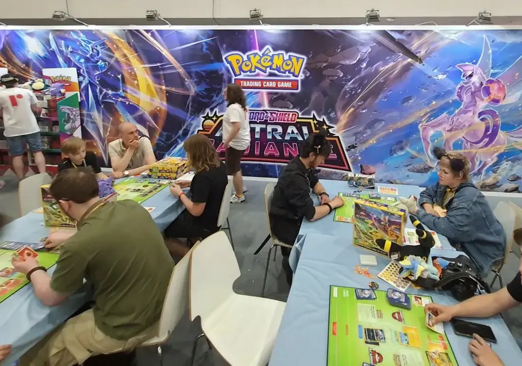 Joueurs de tous âges attablés avec des cartes Pokémon sous une affiche du JCC, au Pyrkon 2022