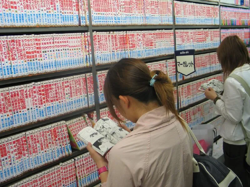 Deux lectrices vues de dos feuillettent des mangas devant des rayonnages de volumes reliés, dans une librairie de Tokyo